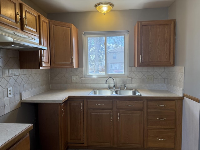 kitchen featuring under cabinet range hood, a sink, light countertops, decorative backsplash, and brown cabinetry