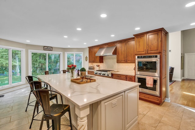 kitchen featuring light stone countertops, range hood, appliances with stainless steel finishes, backsplash, and brown cabinets