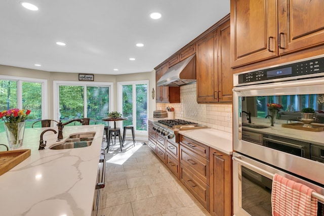 kitchen with brown cabinets, a sink, range hood, stainless steel appliances, and backsplash