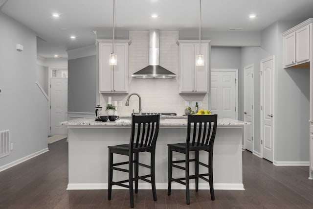 kitchen featuring dark wood-style floors, wall chimney exhaust hood, a center island with sink, and decorative backsplash