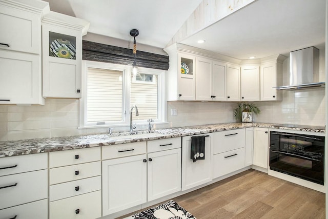 kitchen featuring white cabinets, light wood-style floors, wall chimney range hood, black appliances, and a sink