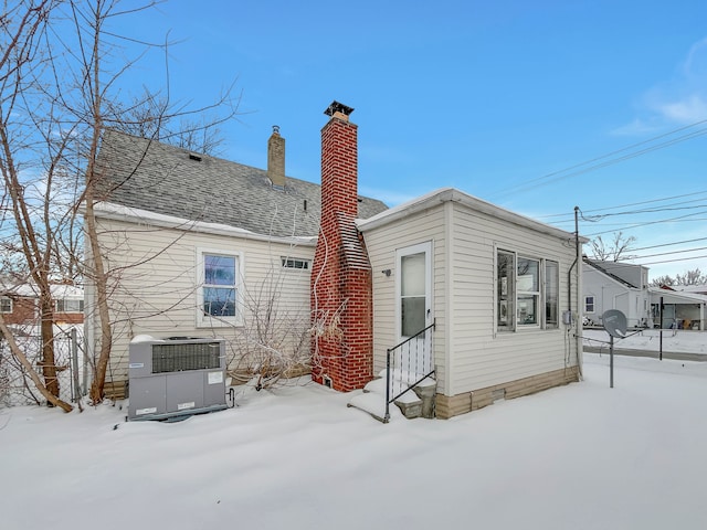 snow covered back of property with entry steps, roof with shingles, central AC unit, and a chimney