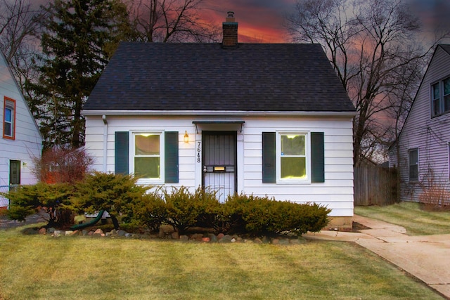 cape cod-style house with a shingled roof, a lawn, a chimney, and fence