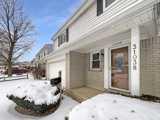 snow covered property entrance featuring brick siding and an attached garage