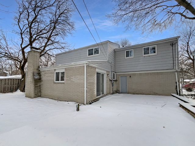 snow covered back of property with a garage, brick siding, a chimney, and fence