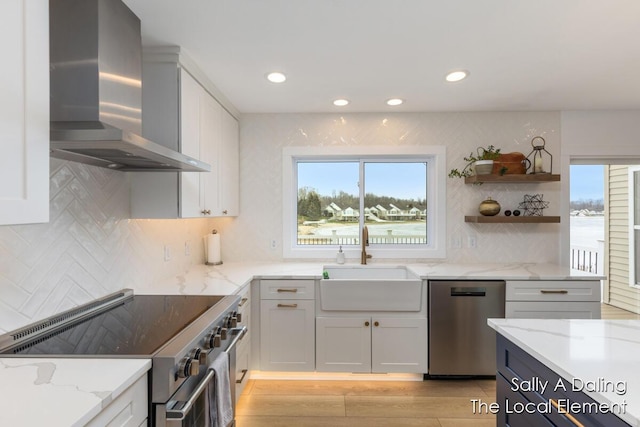 kitchen featuring light stone counters, appliances with stainless steel finishes, white cabinets, a sink, and wall chimney range hood