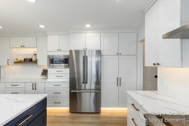 kitchen with light wood finished floors, light stone countertops, range hood, stainless steel appliances, and white cabinetry