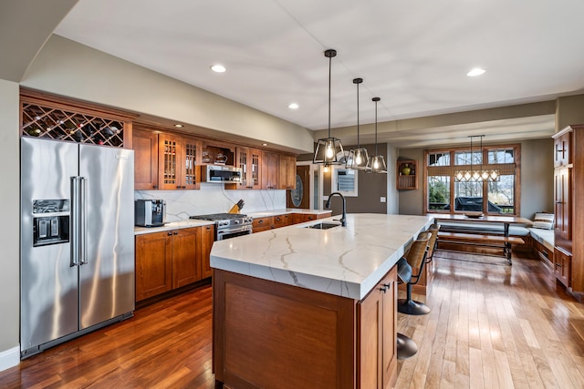 kitchen with a center island with sink, brown cabinetry, high end appliances, light stone countertops, and a sink