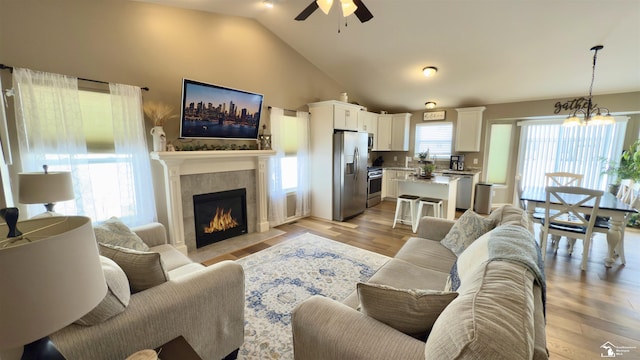 living area featuring ceiling fan with notable chandelier, light wood-type flooring, a tiled fireplace, and high vaulted ceiling