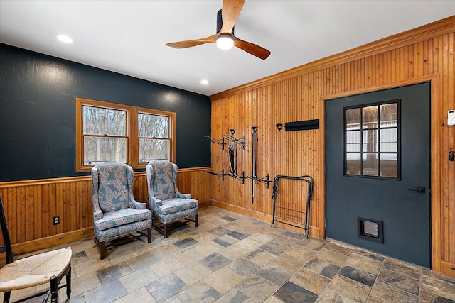 living area featuring wooden walls, a ceiling fan, stone tile flooring, and wainscoting