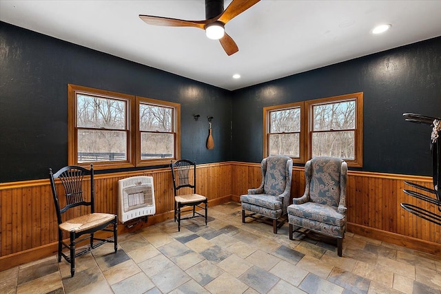living area featuring stone finish flooring, a ceiling fan, heating unit, and wainscoting