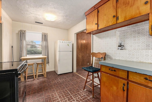 kitchen featuring visible vents, brown cabinetry, freestanding refrigerator, brick floor, and black range with electric cooktop