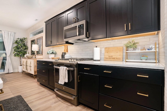 kitchen featuring light wood finished floors, decorative backsplash, stainless steel appliances, dark cabinetry, and light countertops