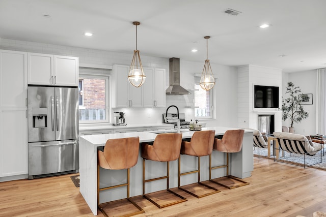 kitchen with a kitchen island with sink, light countertops, wall chimney range hood, stainless steel fridge with ice dispenser, and light wood finished floors