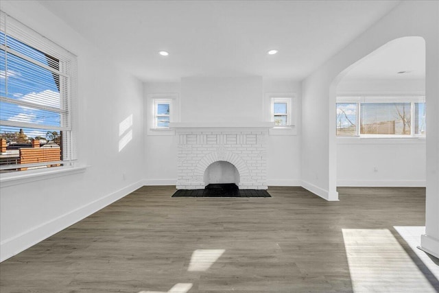 unfurnished living room featuring arched walkways, a brick fireplace, baseboards, and wood finished floors