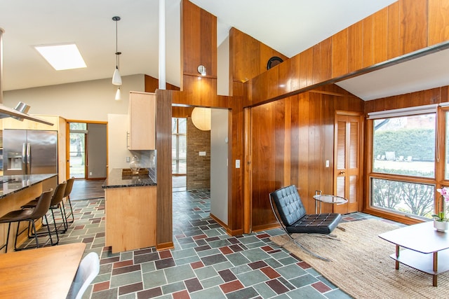 kitchen featuring a skylight, dark stone counters, wood walls, high vaulted ceiling, and stainless steel refrigerator with ice dispenser