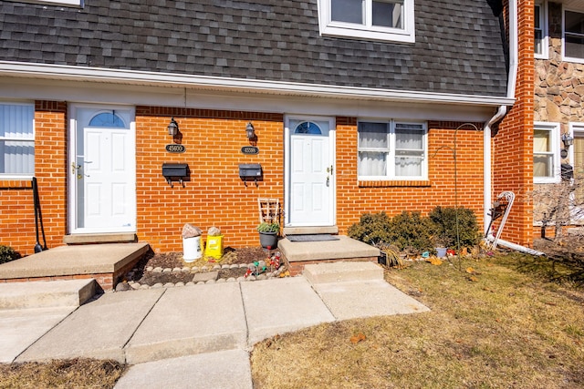 view of exterior entry with brick siding, mansard roof, and a shingled roof