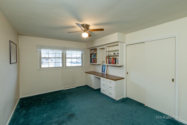 unfurnished office with built in desk, dark colored carpet, baseboard heating, ceiling fan, and baseboards