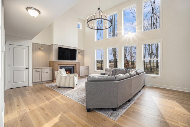 living area featuring a warm lit fireplace, light wood-style flooring, baseboards, and a chandelier