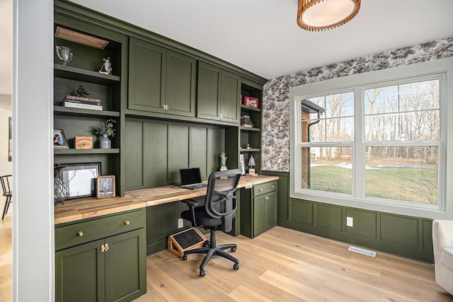 home office with light wood-style flooring, a decorative wall, visible vents, built in desk, and wallpapered walls