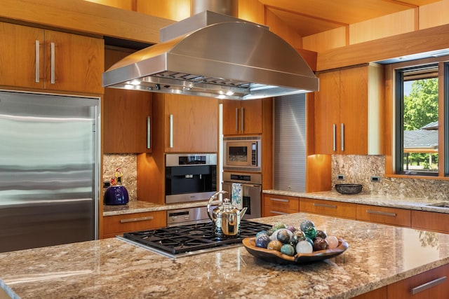 kitchen featuring built in appliances, light stone counters, brown cabinets, and island range hood