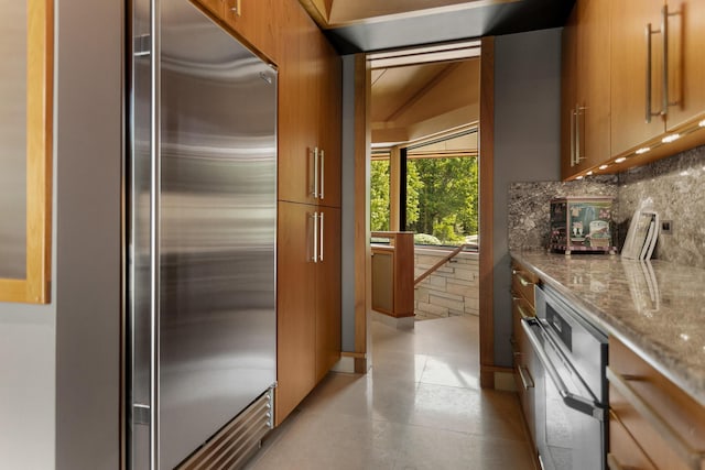 kitchen featuring brown cabinetry, backsplash, built in refrigerator, and light stone countertops