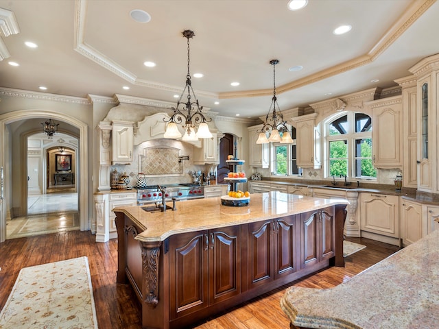 kitchen featuring a tray ceiling, cream cabinets, arched walkways, and an island with sink