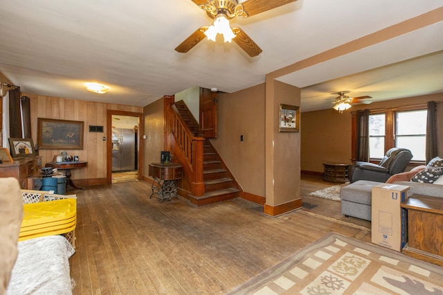 living area featuring stairway, ceiling fan, wood walls, baseboards, and hardwood / wood-style flooring