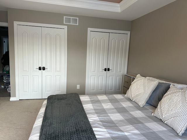 bedroom featuring light carpet, visible vents, a tray ceiling, two closets, and crown molding