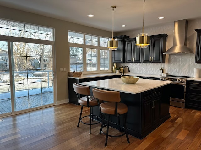 kitchen with wall chimney exhaust hood, dark wood-style flooring, a kitchen island with sink, light countertops, and stainless steel range with electric stovetop