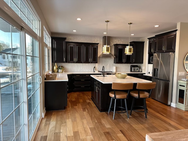 kitchen featuring light wood-style floors, light countertops, wall chimney range hood, stainless steel refrigerator with ice dispenser, and an island with sink