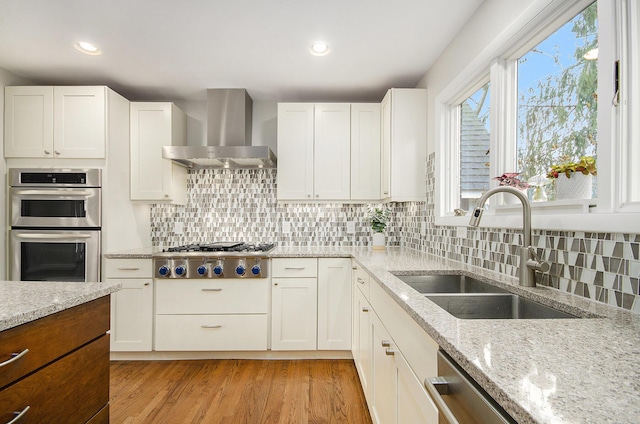 kitchen with appliances with stainless steel finishes, light stone counters, light wood-type flooring, wall chimney range hood, and a sink