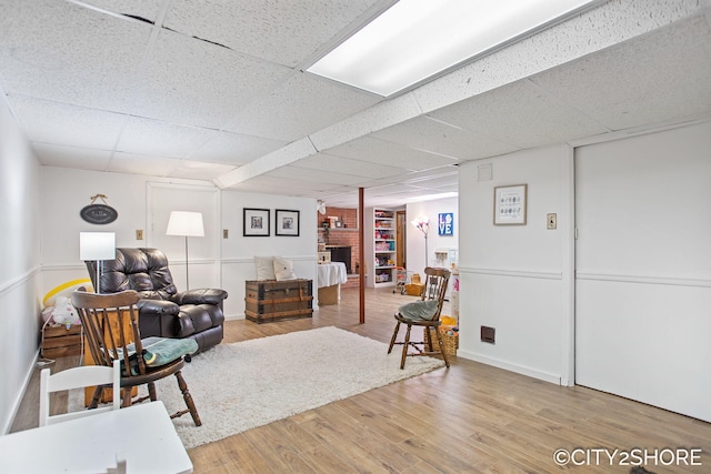 living area with a brick fireplace, a drop ceiling, baseboards, and wood finished floors