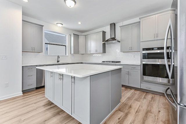 kitchen with gray cabinetry, appliances with stainless steel finishes, a sink, wall chimney range hood, and light wood-type flooring