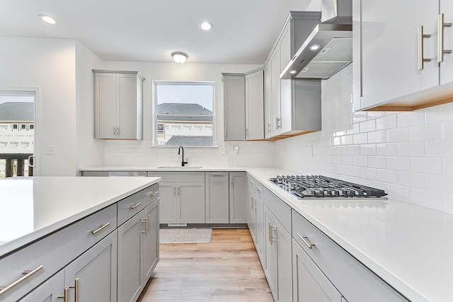 kitchen featuring gray cabinetry, a sink, stainless steel gas cooktop, and wall chimney exhaust hood