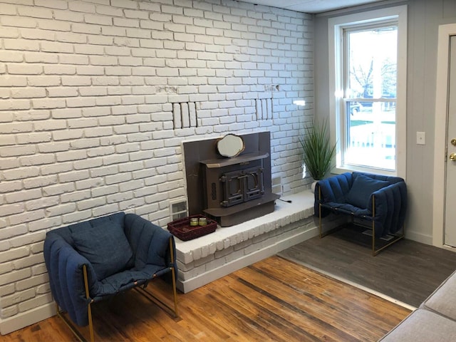 sitting room with a wood stove, a healthy amount of sunlight, brick wall, and wood finished floors