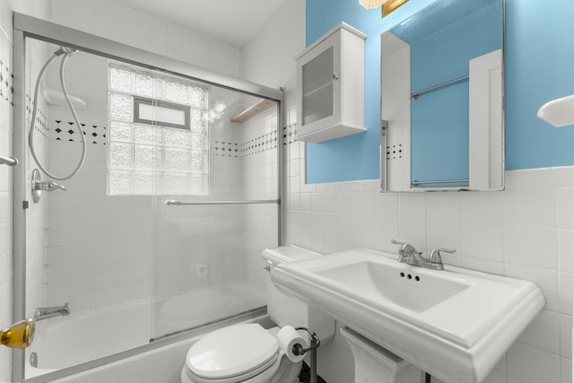 full bathroom featuring toilet, a wainscoted wall, bath / shower combo with glass door, a sink, and tile walls