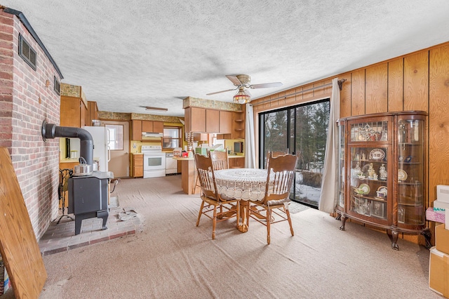 unfurnished dining area featuring light carpet, a ceiling fan, a wood stove, a textured ceiling, and wood walls