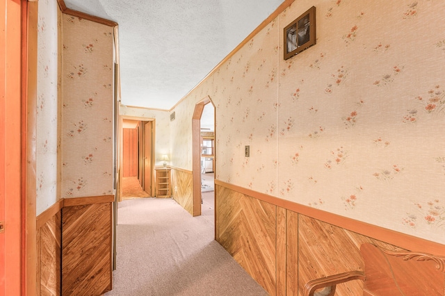 hall featuring wallpapered walls, arched walkways, wainscoting, crown molding, and carpet flooring