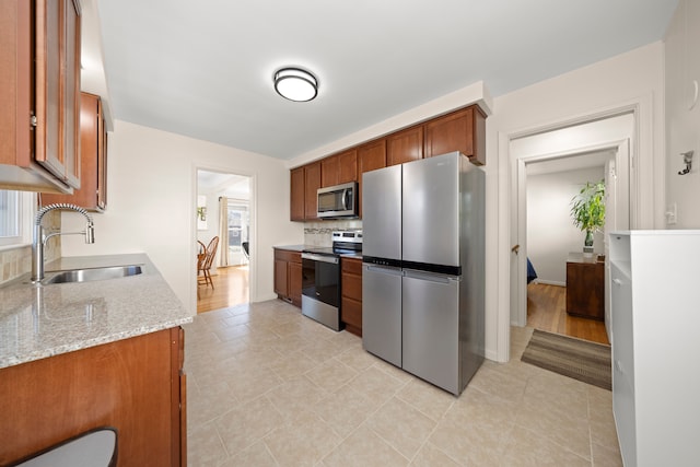 kitchen with brown cabinetry, decorative backsplash, light stone counters, appliances with stainless steel finishes, and a sink