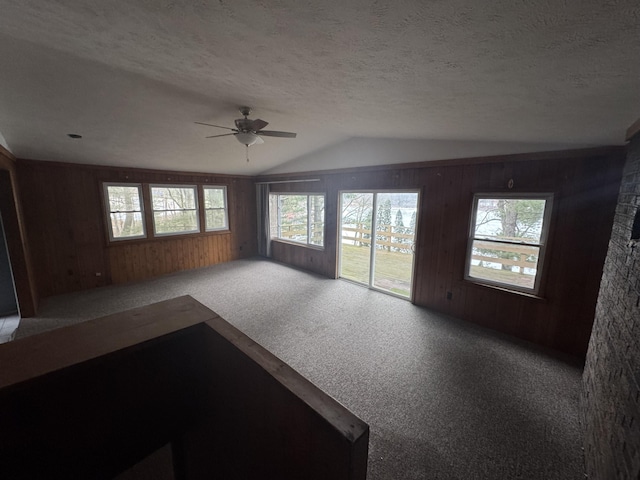 unfurnished living room with a ceiling fan, carpet flooring, vaulted ceiling, wooden walls, and a textured ceiling