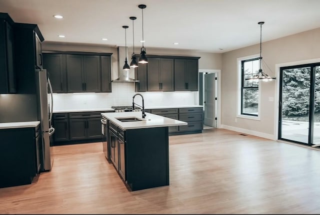 kitchen featuring a sink, light countertops, wall chimney range hood, tasteful backsplash, and a center island with sink