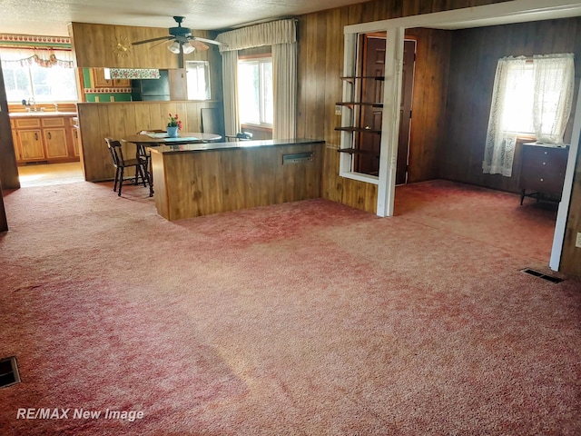 kitchen with light colored carpet, visible vents, a ceiling fan, brown cabinetry, and wood walls