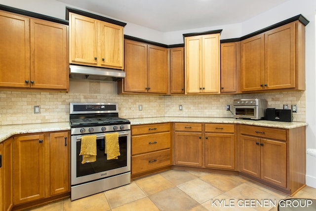 kitchen with light stone countertops, under cabinet range hood, stainless steel gas range oven, and decorative backsplash