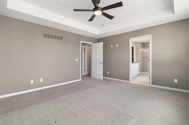 unfurnished bedroom featuring a tray ceiling, light colored carpet, visible vents, ensuite bath, and baseboards