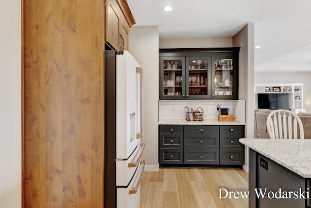 kitchen with tasteful backsplash, white refrigerator with ice dispenser, glass insert cabinets, light stone counters, and light wood-type flooring