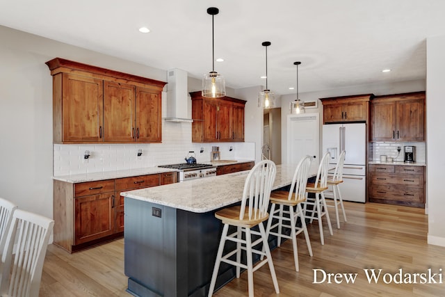 kitchen with white refrigerator with ice dispenser, brown cabinets, light wood finished floors, wall chimney exhaust hood, and a kitchen bar