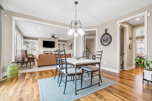 dining area with built in shelves, ceiling fan with notable chandelier, a fireplace, baseboards, and light wood-type flooring