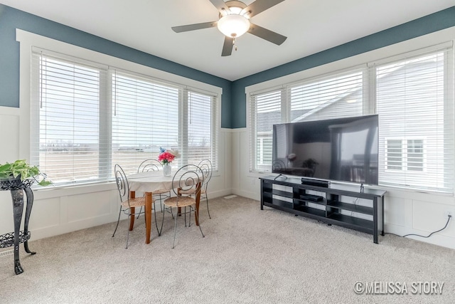 dining space featuring carpet floors, a decorative wall, a ceiling fan, and wainscoting