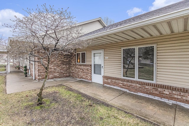 view of exterior entry featuring brick siding, a patio area, and a shingled roof